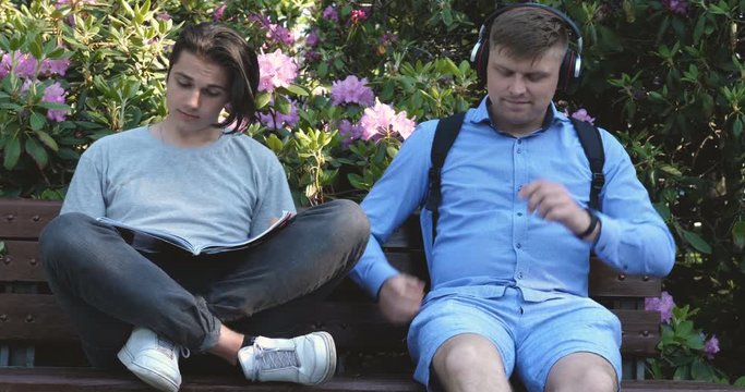 Two beautiful young people Teens, reading a book, listening to music in a mobile phone, sitting on a bench, Background of flowers and growths. Concept: study, students, hairstyles, lifestyle.