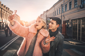 Say cheese. Portrait of cheerful blond girl in pink fur coat taking photo with her boyfriend. They looking at smartphone camera and smiling