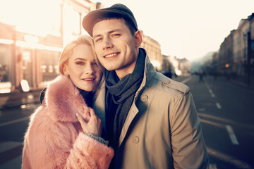 Closer to you. Portrait of stylish guy in flat cap embracing his smiling girlfriend in pink fur coat