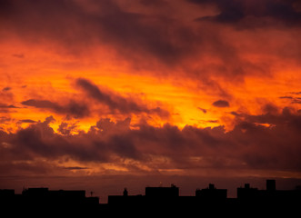 A dramatic and reddish view of a sunset sky with a lot of clouds over a black city silhouette, transmitting a threaten feeling