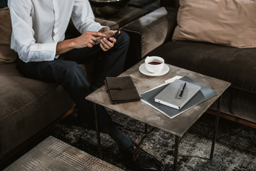 Top view of businessman sitting on couch near end table. He is messaging via smartphone while using day planers for making notes and drinking hot beverage