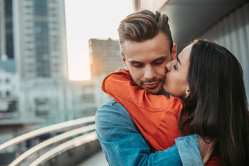 Smiling woman kissing cheerful male in cheek while embracing him outside. Lovers having fun together concept - obrazy, fototapety, plakaty