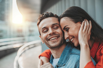 Fototapeta premium Laughing lady talking with cheerful man while hugging him outdoor during leisure. She touching hear with arm