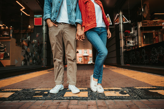 Male holding arm of young woman while locating outdoor near shop-windows. They having fun together while wearing white trainers and standing on road hump