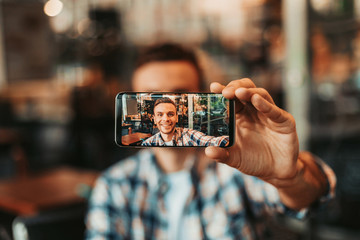 Portrait of positive man taking selfie on mobile while looking at it. Positive guy keeping gadget...