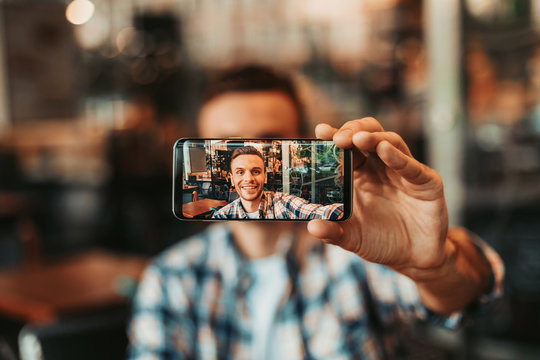 Portrait of positive man taking selfie on mobile while looking at it. Positive guy keeping gadget in hand during fun concept