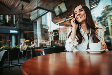 Low angle portrait of happy female talking on phone while locating at desk in cafe. She tasting mug of liquid during conversation with gadget