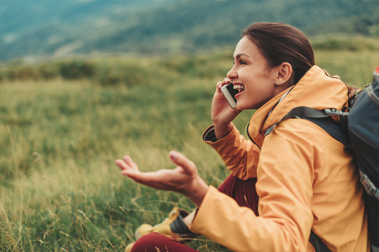 Cheerful delighted young woman sitting in the mountain hills while having a conversation on phone