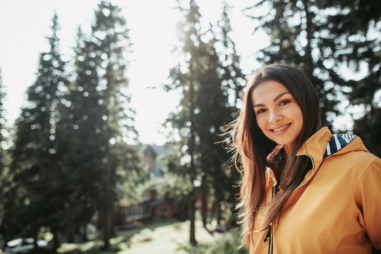 Positive emotions. Close up portrait of beautiful girl in yellow jacket posing in forest on blurred background. She is looking at camera with smile