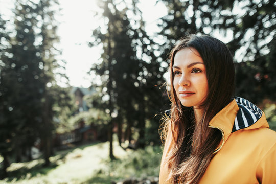 My vacation. Close up portrait of charming girl in yellow jacket standing in forest on blurred background. She is looking away with serious expression