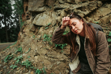 I need some rest. Portrait of tired young lady sitting on hillside and touching forehead with hand. She is having break during long hike