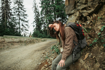 Long way. Portrait of exhausted girl with closed eyes sitting on hillside and touching forehead with hand. She is having break during hike
