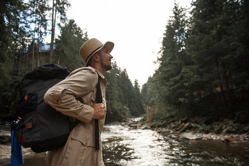 Inspired by journey. Optimistic bearded young man carrying big backpack and looking confident while standing next to the mountain stream and smiling