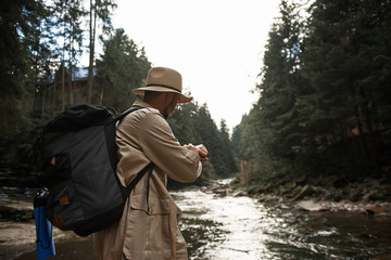 Checking time. Confident experienced traveler carrying his backpack and looking at the watch while standing next to the mountain river