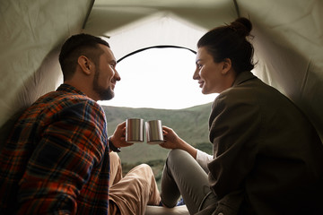 In the tent. Peaceful young couple smiling and enjoying hot tea while sitting in comfortable tent with cups in their hands