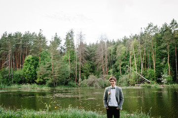 Fototapeta na wymiar The portrait of attractive groom in a suit and bow is standing against the background of the forest at nature near river or lake, pond.