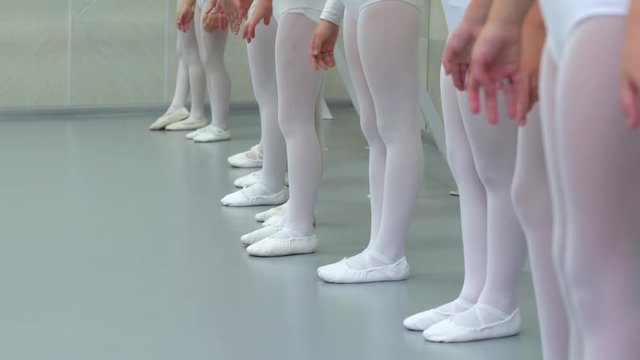 closeup legs of little ballerinas group in white shoes in row practicing in ballet school slow motion. Young girls training poses of classical dance exercise. Childhood, dancing, lifestyle concept