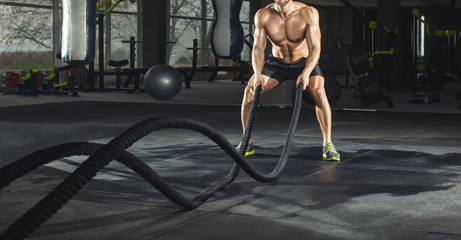 Athletic adult man with battle rope doing exercise in the fitness gym. Sports concept.