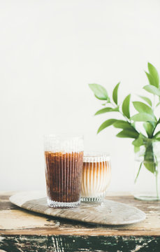 Iced coffee in tall glasses with milk and straws on board over wooden table, white wall at background, copy space. Summer refreshing beverage ice coffee drink concept