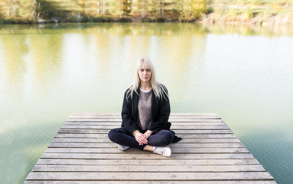 Blonde young woman in black coat sitting in a lotus pose on the wooden pier