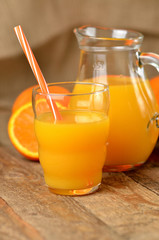 Glass full of orange juice, carafe with fresh juice, straw and fresh oranges in the background on wooden table - vertical photo