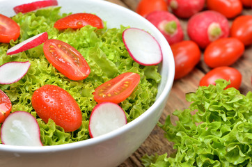 Close-up of fresh green salad Lollo Biondo with wet tomatoes and radishes with drops in a white bowl on wooden table