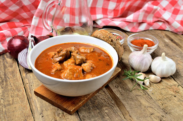 Pork goulash with pieces of meat in a bowl, garlic, pepper, onion, jug with oil and red checkered...