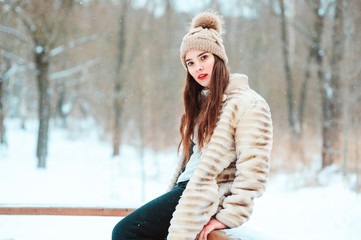 winter portrait of beautiful young woman in fur coat and knitted hat walking in snowy park or...