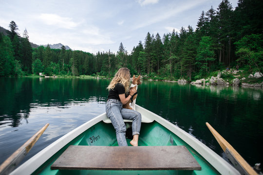 Adorable, cute young woman with blond hair in casual outfit sits together with her best friend, basenji puppy dog and cuddles in rowing boat in lush green forest surroundings of alpine lake