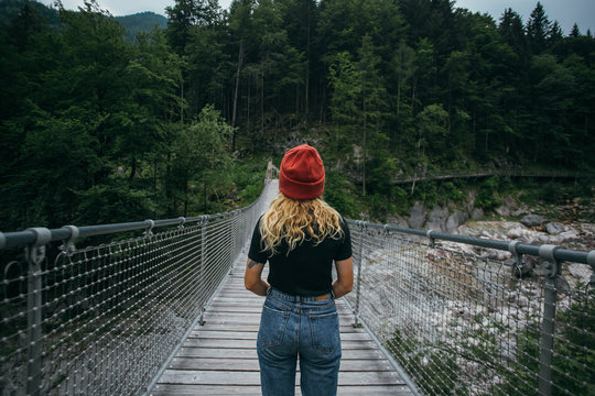 Young explorer, urban nomad and female adventurer in middle of hanging wooden authentic bridge in forest mountains, wears millennial hipster outfit for hiking or trekking, wanderlust concept lifestyle