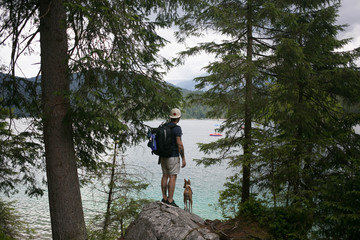 Young adventurous man with backpack and travel companion little puppy dog, best friend explore forest nature in mountains together.Stand in trees next to lake, tourism in rural aread,camping lifestyle