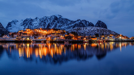Lofoten islands, Norway. Colorful winter landscape in blue hours. Illuminated fishing village reflected in water. Snowy mountains in background. 