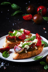 Crispy bruschetta with red and brown cherry tomatoes, feta cheese and green basil on the plate, selective focus