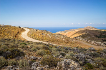 Empty road cutting through yellow hill with a sea in the distance, Crete, Greece