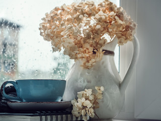 Cup of strong coffee, bouquet of beautiful, white flowers in a vintage jug on the background of a rainy, cloudy day
