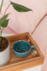 Green monstera plant on the wooden tray on the nightstand. Big blue empty cup on the tray on the coffee table at the bed. Pink bedding