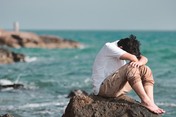 Anxious confused Asian man covering face with hands and bend down head on the rock of sea shore.