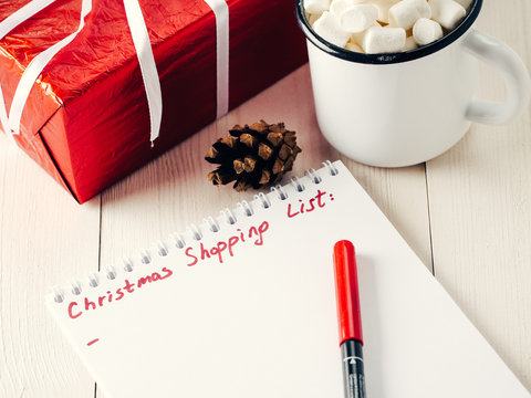 Christmas gifts shopping planning. Make shopping or to-do list for Christmas. Notebook, mug hot chocolate with marshmallows, New Year's gift and pine cone on white wooden background.