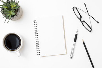 Blank notebook is on top of white office desk table with coffee cup and office supplies. Top view with copy space, flat lay.