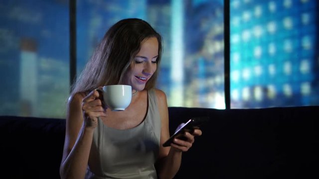 Young woman messaging at home using smartphone, relaxing, drinking tea at home, modern new flat