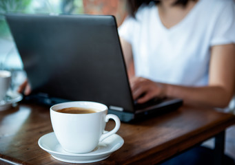 Coffee cup on wooden table with blurred girl using laptop.