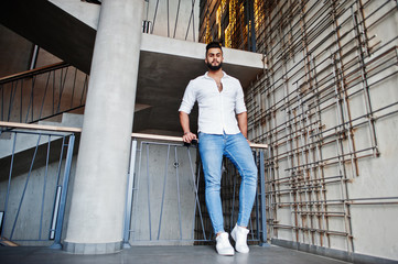 Stylish tall arabian man model in white shirt, jeans posed against steel wall indoor. Beard attractive arab guy.