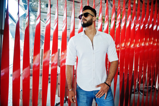 Stylish tall arabian man model in white shirt, jeans and sunglasses posed at street of city. Beard attractive arab guy against red wall.