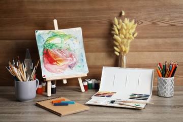 Easel with abstract painting and set of professional art supplies on table against wooden background