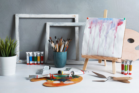 Easel with abstract painting and set of professional art supplies on table against grey background