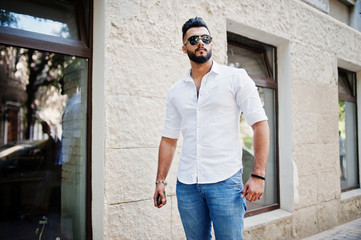 Obraz na płótnie Canvas Stylish tall arabian man model in white shirt, jeans and sunglasses posed at street of city. Beard attractive arab guy.