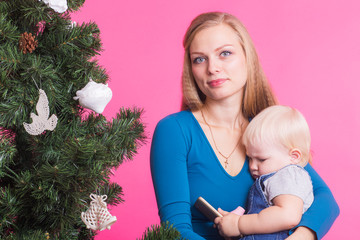 Holidays, family and christmas concept - young woman with her baby near christmas tree on pink background