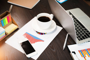 White ceramic coffee cup and mobile phone put on business paper chart,beside laptop,on wooden desk,at office,
