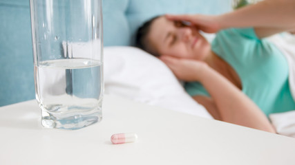 Obraz na płótnie Canvas Closeup image of pill and glass of water on bedside table against sick woman lying in bed