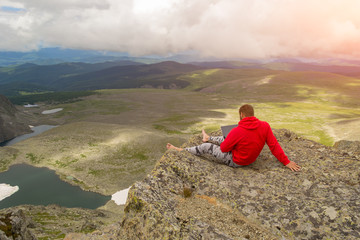 A young man sits barefoot with a laptop in his hands work as a freelancer while traveling on the edge of a high mountain on a dangerous cliff at the bottom of which the lake and field with plants